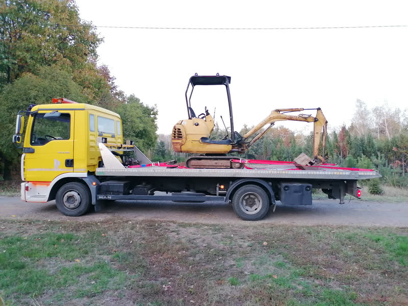 Ein gelber MAN-Abschleppwagen von der Seite, mit einem Minibagger, der auf dem Plateau festgezurrt ist.