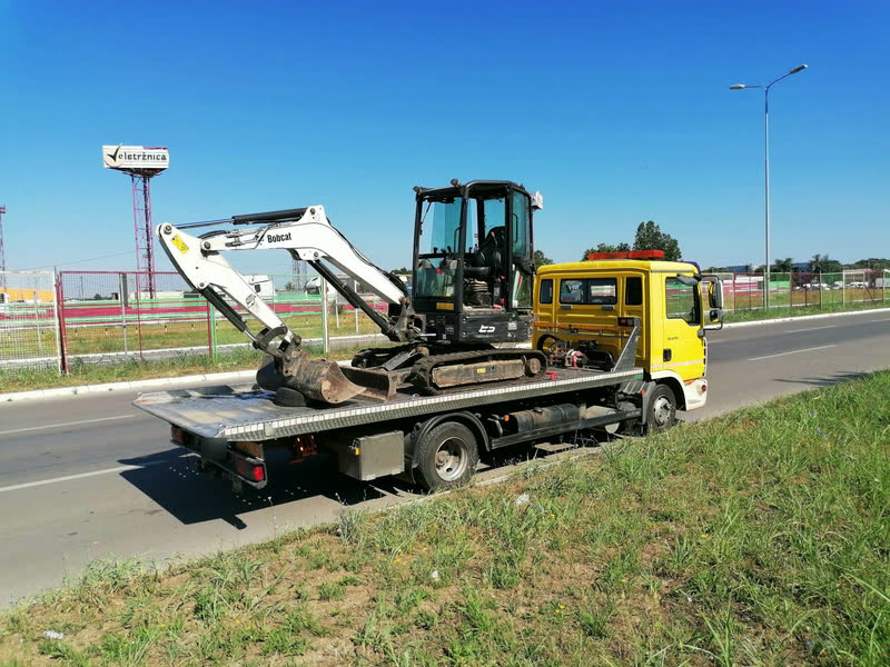 Ein weißer Bobcat-Bagger auf dem Plateau eines gelben MAN-Abschleppwagens am Straßenrand, an einem sonnigen Tag.