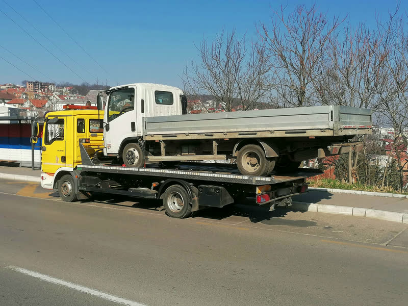 Ein kleiner weißer Lkw mit Aluminiumpritsche auf dem Plateau eines gelben MAN-Abschleppwagens.